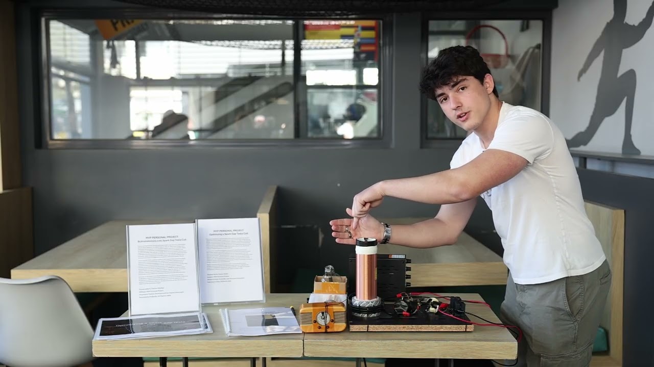 Paul presenting his Tesla coil prototype on a table: a copper-wound secondary coil on a wooden base with a driver circuit, beside printed project posters and the written report.