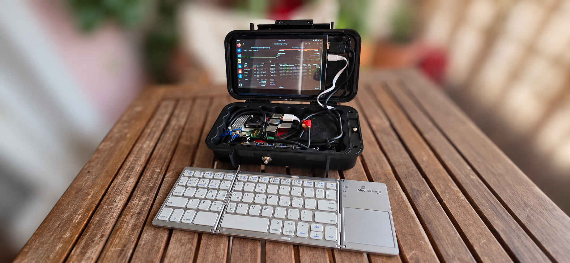 Paul's homemade laptop: a black hard shell case open on a wooden table, with a 7-inch screen in the lid showing an Ubuntu desktop and system monitor, a Raspberry Pi 5 with a finned heatsink and fan in the base, and a silver tri-fold keyboard with trackpad in front.